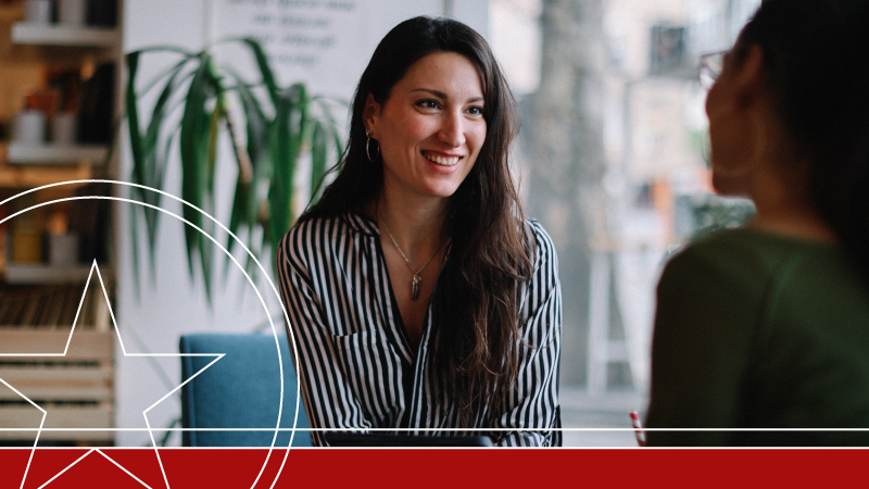 Woman in striped business shirt smiling in modern office setting with plant in background.
