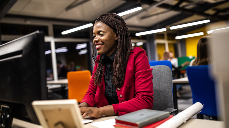Young cheerful African woman reading some paper documents while sitting at desk in the office, using computer