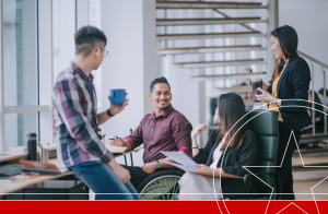 Indian white collar male worker in wheelchair having cheerful discussion leading conversation with colleague in creative office workstation beside window
